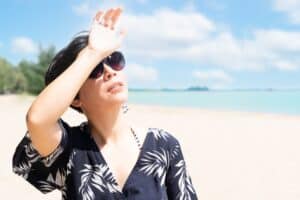 Woman standing on the beach holding a hand up to block her face from the sun and in need of skin protection.